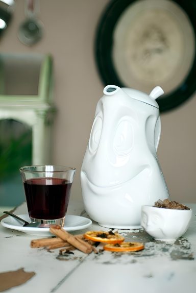 Weiße Kanne, Glas mit rotem Getränk, Zimtstangen und trockenen Orangenscheiben auf Tisch.